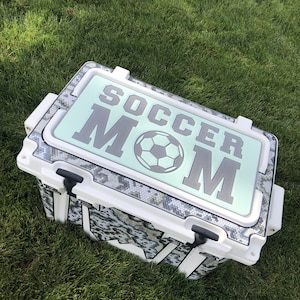 May include: White cooler with a black and gray snake skin pattern. The lid has a light green background with the words "Soccer Mom" in gray and a black soccer ball.
