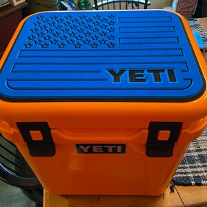 Puede incluir: Una nevera YETI naranja con una tapa azul con un dise&ntilde;o de bandera estadounidense negra. La nevera tiene cierres negros y el logotipo de YETI en la parte delantera. Se ve un asa azul.