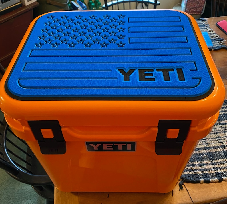 Puede incluir: Una nevera YETI naranja con una tapa azul con un dise&ntilde;o de bandera estadounidense negra. La nevera tiene cierres negros y el logotipo de YETI en la parte delantera. Se ve un asa azul.