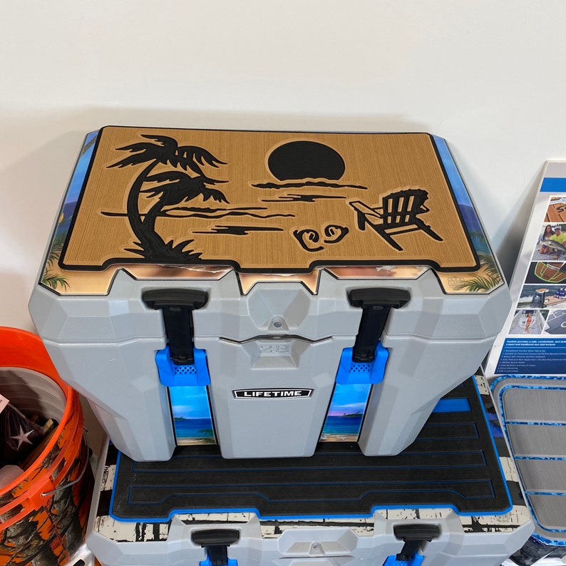 May include: A gray cooler with a brown lid featuring a black silhouette of a palm tree, a sun, and a beach chair. The cooler has blue latches and the word "LIFETIME" printed on the front.