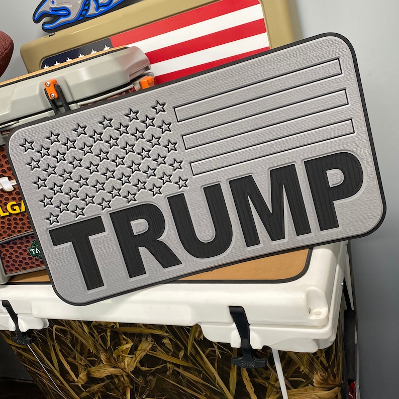 May include: A rectangular, silver-colored sign with a textured surface. The sign features a design of the American flag with the word "TRUMP" in bold, black letters. The sign is resting on top of a white cooler.