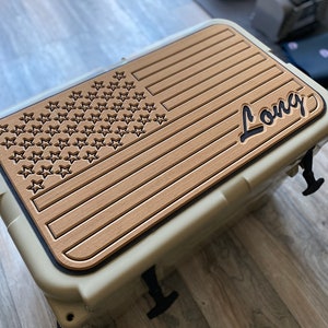 K&ouml;nnte beinhalten: Beige K&uuml;hler mit einem holzstrukturierten Deckel mit einem eingravierten amerikanischen Flaggen-Design und dem Namen "Long" in einer Schreibschrift. Der K&uuml;hler hat schwarze Verschl&uuml;sse und einen Griff. Der K&uuml;hler steht auf einem Holzboden.