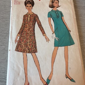 May include: A Simplicity 7194 sewing pattern envelope featuring illustrations of two dresses. One is a long-sleeved dress with a paisley print and a solid collar, and the other is a short-sleeved solid teal dress with a buttoned placket and collar.