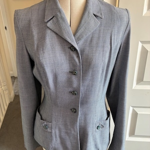 May include: A blue and white micro-check blazer featuring a notched lapel, long sleeves, and a front button closure with four matching buttons. The garment includes two front patch pockets, each accented with a single button, and structured shoulders.