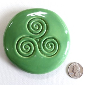 Puede incluir: Una pieza de cer&aacute;mica redonda y brillante, de color verde claro, con un dise&ntilde;o de triple espiral en relieve. Una moneda est&aacute; al lado para la comparaci&oacute;n de tama&ntilde;o. El dise&ntilde;o es un s&iacute;mbolo celta.