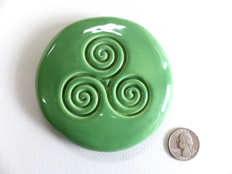 Puede incluir: Una pieza de cer&aacute;mica redonda y brillante, de color verde claro, con un dise&ntilde;o de triple espiral en relieve. Una moneda est&aacute; al lado para la comparaci&oacute;n de tama&ntilde;o. El dise&ntilde;o es un s&iacute;mbolo celta.