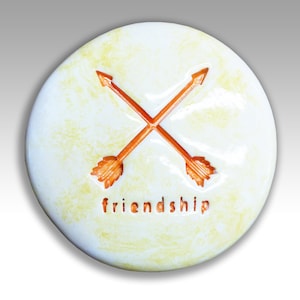May include: A white ceramic coaster with a yellow glaze and two orange arrows crossed in the center. The word "friendship" is written below the arrows.