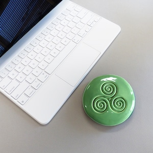 Puede incluir: Un posavasos de cer&aacute;mica verde claro con un dise&ntilde;o de espirales celtas se encuentra junto a un port&aacute;til blanco sobre una superficie gris. El posavasos es redondo y tiene un acabado brillante. El teclado del port&aacute;til es visible.