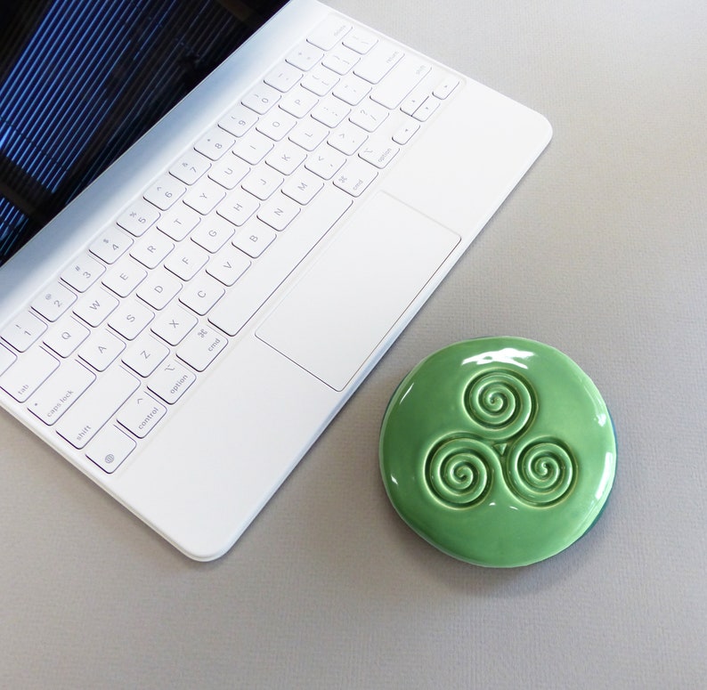 Puede incluir: Un posavasos de cer&aacute;mica verde claro con un dise&ntilde;o de espirales celtas se encuentra junto a un port&aacute;til blanco sobre una superficie gris. El posavasos es redondo y tiene un acabado brillante. El teclado del port&aacute;til es visible.