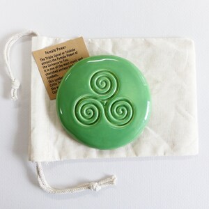Puede incluir: Una pieza redonda de cer&aacute;mica verde claro brillante con un dise&ntilde;o de triple espiral en relieve, colocada sobre una bolsa de cord&oacute;n crema. Una peque&ntilde;a etiqueta con texto sobre el "Poder Femenino" es visible. El dise&ntilde;o es un s&iacute;mbolo celta.