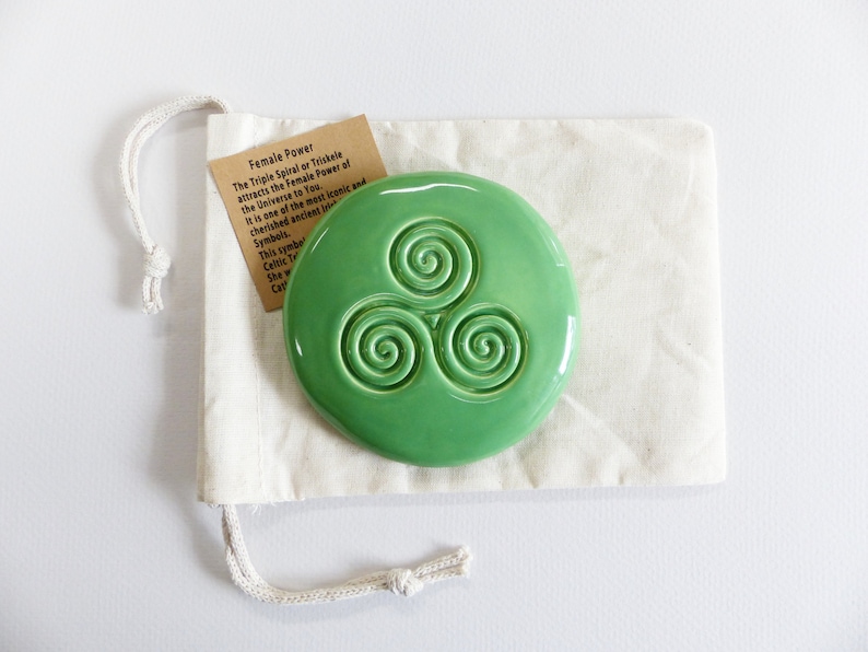 Puede incluir: Una pieza redonda de cer&aacute;mica verde claro brillante con un dise&ntilde;o de triple espiral en relieve, colocada sobre una bolsa de cord&oacute;n crema. Una peque&ntilde;a etiqueta con texto sobre el "Poder Femenino" es visible. El dise&ntilde;o es un s&iacute;mbolo celta.