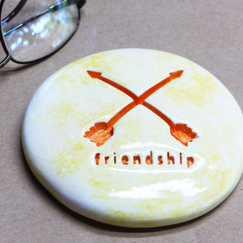 May include: White ceramic coaster with a yellow glaze. The coaster has a design of two orange arrows crossing each other. The word "friendship" is written below the arrows.