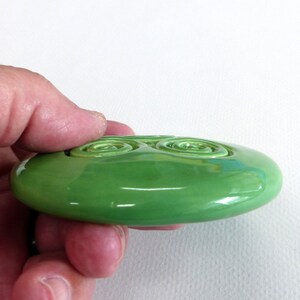 Puede incluir: Una pieza de arte de vidrio verde brillante de forma ovalada. La superficie presenta dos dise&ntilde;os en espiral. La pieza se sostiene en una mano sobre un fondo blanco. El vidrio tiene una calidad reflectante.