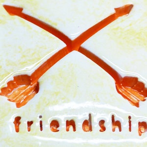 May include: A white ceramic tile with two orange arrows crossing each other. The word "friendship" is written in a cutout font below the arrows.