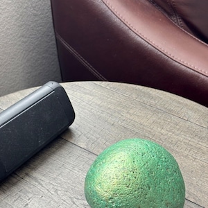 May include: A rounded, textured stone with a metallic, iridescent green finish that shifts in color. The surface of the stone is uneven and porous, reflecting light across its curved shape, creating a distinct shimmer against the surrounding area.