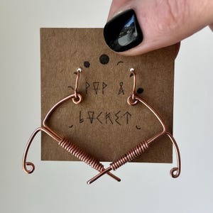 May include: A pair of copper wire earrings shaped like hearts with a twisted wire design. The earrings are displayed on a brown card with the text "POP & LOCKET" written on it.