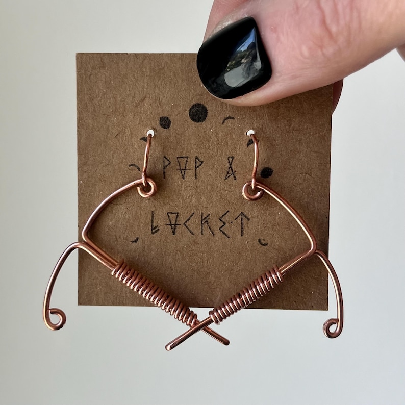May include: A pair of copper wire earrings shaped like hearts with a twisted wire design. The earrings are displayed on a brown card with the text "POP & LOCKET" written on it.