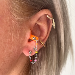 May include: A close-up of a person's ear with multiple piercings. The ear is pierced with a hoop earring, a small cross earring, a lightning bolt earring, and a chain with colorful beads. The chain is attached to a small orange earplug.