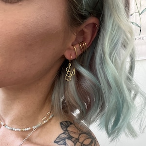 May include: Gold hoop earrings with a gold initial charm hanging from the bottom. The earrings are worn in a person's ear with other gold hoop earrings.