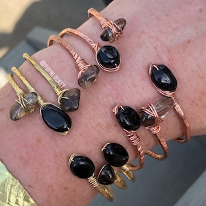 Könnte beinhalten: Vier goldene und kupferne, drahtgewickelte Armbänder mit schwarzen und braunen Edelstein-Akzenten. Die Armbänder sind auf dem Handgelenk einer Person gestapelt. Der Text '@POPNLOCKET' ist auf einem der Armbänder sichtbar.