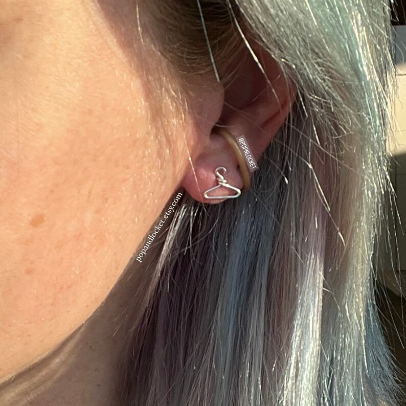 May include: Silver hanger earrings. The earrings are small and delicate, and they are shaped like a hanger. The earrings are worn by a person with grey hair. The earrings are a popular and trendy accessory.