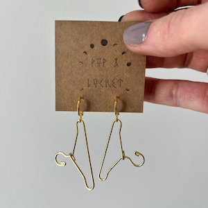 May include: Gold-toned hanger earrings with a hoop design. The earrings are displayed on a brown card with the text "POP & LOCKET" written on it.