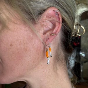 May include: A pair of dangle earrings featuring a unique design. Each earring has a silver hook and a charm shaped like a cigarette, with orange, white, and black details. The earrings are worn on a person's ear.