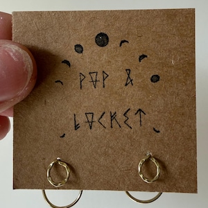 May include: A pair of gold-colored hoop earrings with a hammered texture, displayed on a brown card. The card has the words "POP & LOCKET" written on it, along with a crescent moon design.