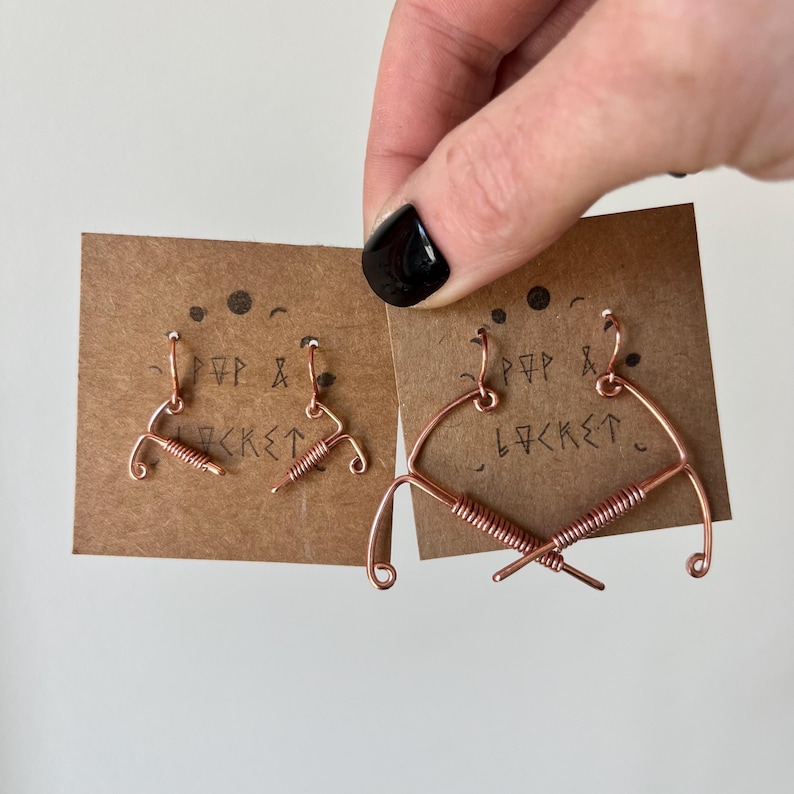 May include: Two pairs of copper wire earrings, one pair is a simple bow shape and the other pair is a more elaborate bow shape with a twisted wire detail. The earrings are displayed on a brown card with the text "FPP & LOCKET" written on it.