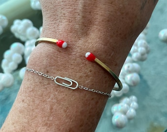 May include: A gold bangle bracelet with red and white accents. The bracelet is worn on a person's wrist. A silver chain bracelet with a paperclip charm is also worn on the wrist.