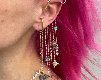 May include: A gold ear cuff with a silver hoop and a long chain with multiple small beads and charms hanging from it. The charms include a star, a blue and white bead, a red bead, a brown bead, a white bead, and a gold bead with a ring around it.