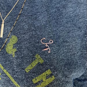 May include: A close-up of a dark gray t-shirt with a gold and black necklace and a small, rose gold wire sculpture of a syringe. The t-shirt has a green graphic print of the word "THE" on it.