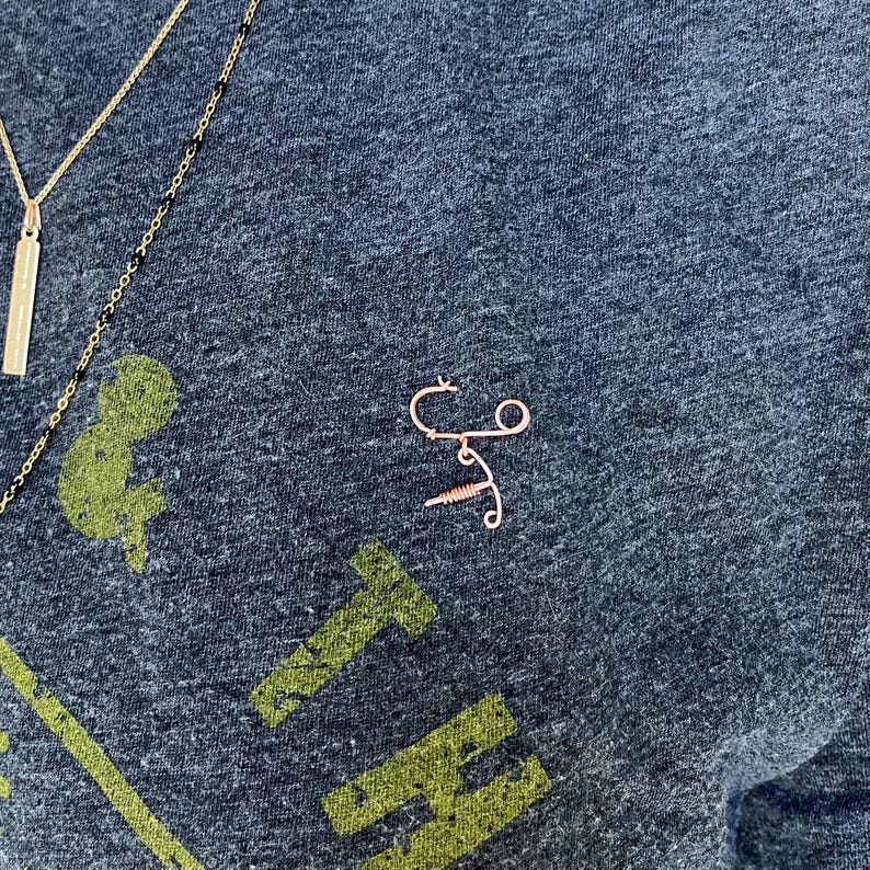 May include: A close-up of a dark gray t-shirt with a gold and black necklace and a small, rose gold wire sculpture of a syringe. The t-shirt has a green graphic print of the word "THE" on it.