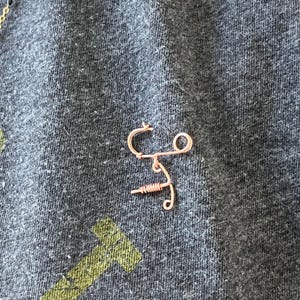 May include: A copper wire sculpture of a stylized figure with a long, thin body and a circular head. The sculpture is on a dark gray fabric background.