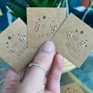May include: A hand holding three pairs of paperclip-shaped stud earrings in silver, rose gold, and gold colors. Each pair is mounted on a small, square, brown card with the text "P & LOCKET" and a small face design.