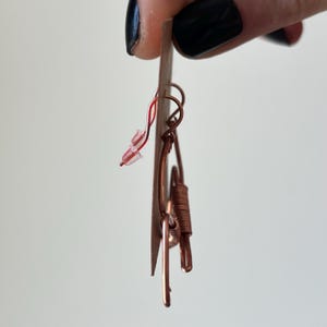 May include: A pair of copper wire earrings with a unique design. The earrings are on a white card.