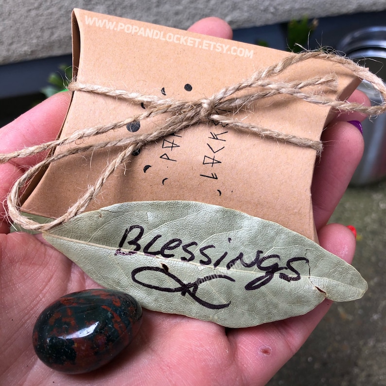 May include: A small, brown, paper box tied with twine and labeled "www.popandlocket.etsy.com". The box is sitting on a hand holding a green leaf with the word "Blessings" written on it. A red and green stone is also in the hand.