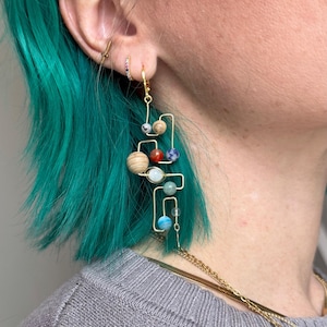 Può includere: Orecchini pendenti dorati con un design geometrico che presenta perline colorate che rappresentano i pianeti. Gli orecchini sono indossati da una persona con i capelli blu turchese.