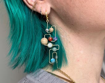 May include: Gold dangle earrings with a geometric design featuring colourful beads representing planets. The earrings are worn by a person with teal blue hair.