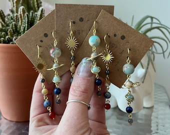 May include: Four pairs of gold solar system earrings with different coloured beads and planets. The earrings are displayed on brown cards with the text "PEP & VIBES".