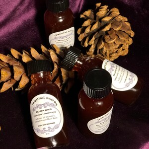 May include: Four small glass bottles with black caps filled with a dark red liquid. The bottles are labeled "Spiritual Magi Oil" and "Dragon Blood" with the text "Purgative Money Love" and "Safflower oil, Vitamin E, Infused Dragon Blood Resin" and "Every Witchcorner" on the label. The bottles are sitting on a purple velvet surface with pine cones.