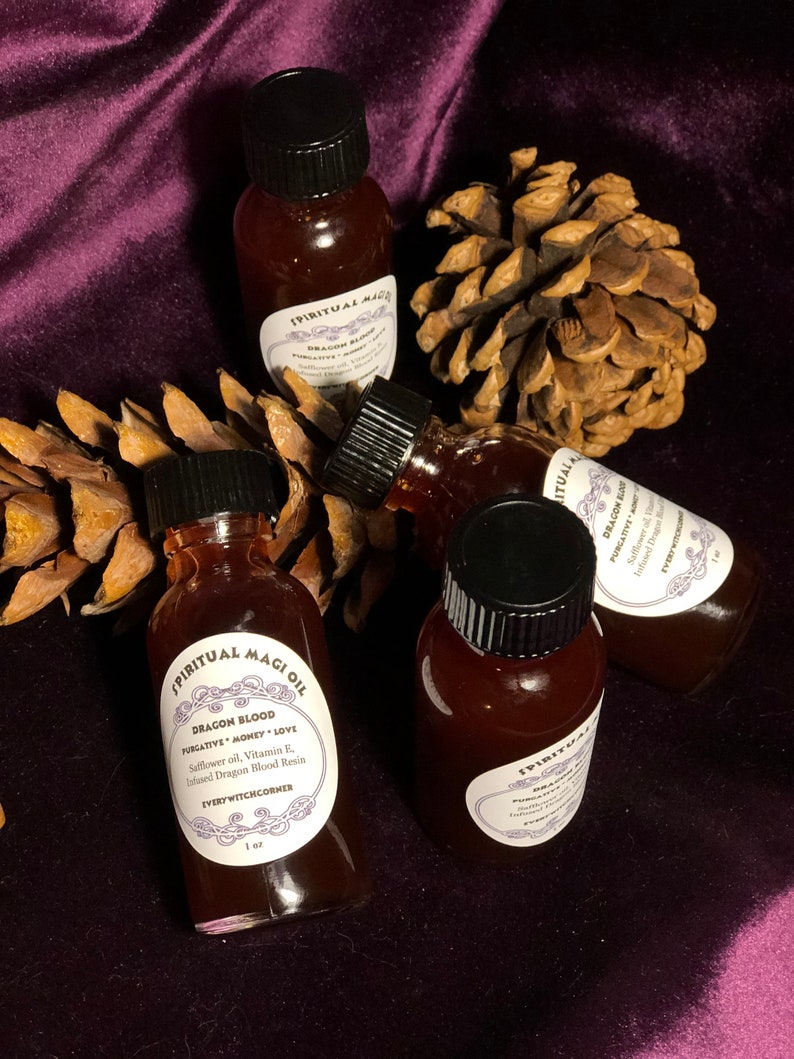May include: Four small glass bottles with black caps filled with a dark red liquid. The bottles are labeled "Spiritual Magi Oil" and "Dragon Blood" with the text "Purgative Money Love" and "Safflower oil, Vitamin E, Infused Dragon Blood Resin" and "Every Witchcorner" on the label. The bottles are sitting on a purple velvet surface with pine cones.