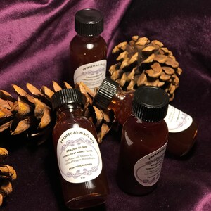 May include: Four small glass bottles with brown liquid and black lids. The bottles are labeled "SPIRITUAL MAGIOIL" and "DRAGON BLOOD PURGATIVE MONEY LOVE" with the text "Safflower oil, Vitamin E, Infused Dragon Blood Resin" and "EVERY WITCHCORNER 1 oz". The bottles are sitting on a brown pine cone and a purple velvet surface.