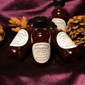 May include: Six small bottles of dark red liquid with black lids and white labels. The labels say "Spiritual Magick Oil" and "Dragon Blood" with the words "Purgative, Money, Love" underneath. The bottles are on a purple velvet surface.