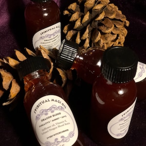 May include: Three small glass bottles filled with a dark red liquid. The bottles have black caps and white labels with the text "SPIRITUAL MAGIOIL DRAGON BLOOD PURGATIVE MONEY LOVE Safflower oil, Vitamin E, Infused Dragon Blood Resin EVERY WITCHCORNER 1 oz" printed on them.