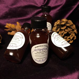 May include: Three small glass bottles with black lids and labels. The labels are white with black text and a decorative border. The bottles contain a dark red liquid. The labels read "Spiritual Magic Oil", "Dragon Blood", "Purgative Money Love", "Safflower oil, Vitamin E, Infused Dragon Blood Resin", and "Every Witch Corner".