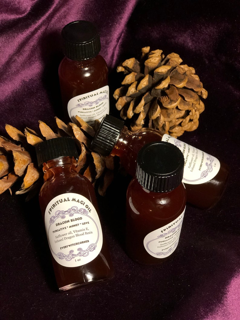 May include: Four small glass bottles filled with a dark red liquid. The bottles have black caps and white labels with the text "SPIRITUAL MAGI OIL" and "DRAGON BLOOD" on them. The bottles are sitting on a brown pine cone and a purple velvet surface.