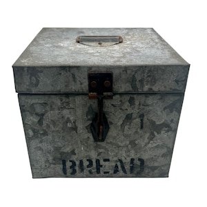 Może przedstawiać: Ocynkowany metalowy pojemnik na chleb z klapą na zawiasach i uchwytem. Pudełko jest prostokątne o lekko trapezowym kształcie i ma słowo "BREAD" na froncie w kolorze czarnym. Posiada rustykalną, industrialną estetykę.