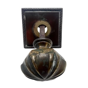 May include: An antique bronze door pull, featuring a square backplate with a rope-style border. The knob is ridged and bulbous. The metal has a weathered appearance. The door pull is likely made of bronze or a similar metal.