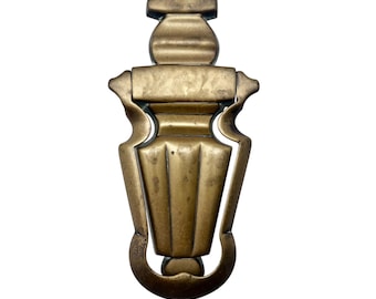 May include: A vintage brass door knocker with an ornate design. The knocker features a central, fluted body, a decorative top, and a curved bottom. The brass has a warm, aged patina, suggesting its antique nature. The overall design is symmetrical and elegant.